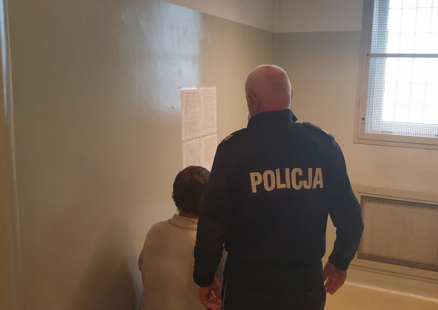 Na zdjęciu mężczyzna siedzący w policyjnej celi, a za min stojący policjant w mundurze.