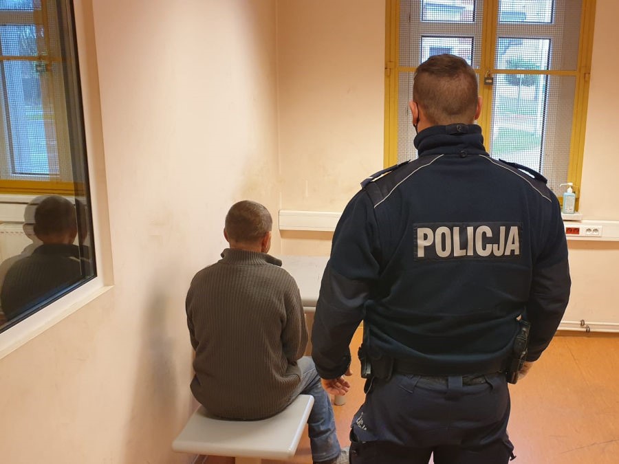 Na zdjęciu policjant stojący za zatrzymanym mężczyzną w pomieszczeniu dla zatrzymanych.