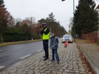 Na zdjęciu umundurowany policjant trzyma radar do mierzenia prędkości obok stoi chłopiec  chłopiec w niebieskiej kurtce.