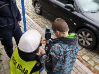 Na zdjęciu umundurowana policjantka pokazuje urządzenie do mierzenia prędkości chłopcu ubranemu w niebieska kurtkę.