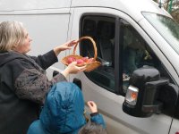 Na zdjęciu kobieta z koszykiem w ręku częstuje kierowcę białego busa jabłkami przez otwarte okno pojazdu.