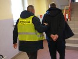 Na zdjęciu policjant w kamizelce  prowadzi korytarzem mężczyznę oskarżonego o usiłowanie zabójstwa dziecka, który ma skute  ręce z tyłu kajdankami.