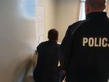 Na zdjęciu zatrzymany mężczyzna w policyjnym areszcie i stojący za nim policjant w mundurze.