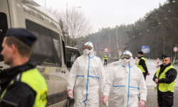 Na zdjęciu policjant w czasie kontroli drogowej w tle widoczne osoby w kombinezonach ochronnych.