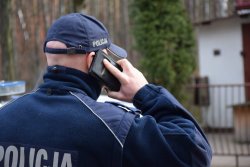 Na zdjęciu umundurowany policjant rozmawiający przez telefon komórkowy trzymany w ręce.