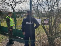 Na zdjęciu policjant z żołnierzem stojący przed furtką ogródków działkowych.