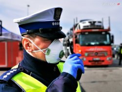 Na zdjęciu policjant w maseczce stojący na tle samochodów  ciężarowych