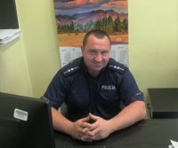 Na zdjęciu mężczyzna w policyjnym mundurze siedzący za biurkiem , dzielnicowy gminy Woźniki asp. Adam Blacha.