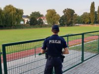 Na zdjęciu widoczny policjant w mundurze stojący tyłem do objetywu na tle boiska sportowego.