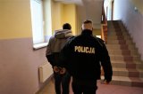 Na zdęciu policjant prowadzi zatrzymanego mężczyznę skutego kajdankami.