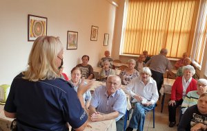 Na zdjęciu policjantka w mundurze w trakcie prelekcji dla Seniorów stojąca tyłem do obietywu a na drugim planie widzoczni uczestnicy spotkania .