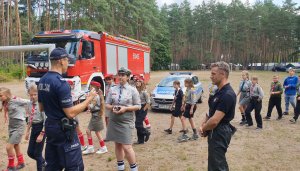 Na zdjęciu policjant w trakcie kontroli obozu harcerskiego w Kokotku rozmawia z kobietą w mundurze harcerskim obok stoi strażak.