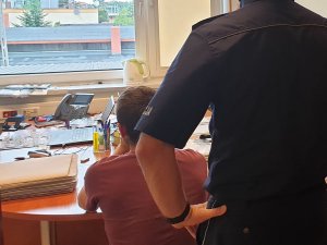 Na zdjęciu zatrzymany mężczyzna siedzący przy stoliku a za nim policjant w mundurze stojący tyłem do obiektywu.