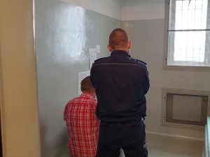 Na zdjęciu policjant w mundurze stojący za siedzacym w celi mężczyzną.