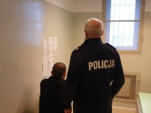 Na zdjęciu mężczyzna siedzący w policyjnej celi, a za nim stojący policjant.