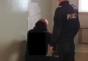 Na zdjeciu widoczna osoba w policyjnej celi , a obok stoi umundurowany policjant.