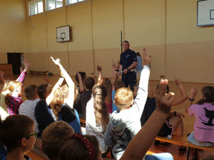 Na zdjęciu policjant w trakcie prelekcji dla dzieci, dzieci zgłaszają się chcąc udzielić odpowiedzi na zadane pytanie podnosząc ręce do góry.
