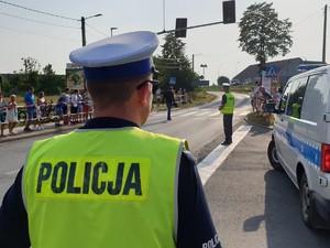 Na zdjęciu policjant  w trakcie zabezpieczenia trasy wyścigu kolarskiego w Woźnikach kierujący ruchem na drodze. W tle widoczny radiowóz policyjny.