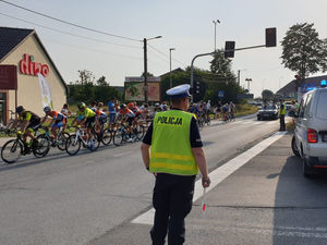 Na zdjęciu policjant  w trakcie zabezpieczenia trasy wyścigu kolarskiego w Woźnikach kierujący ruchem na drodze. W tle widoczni przejeżdżający kolarze.