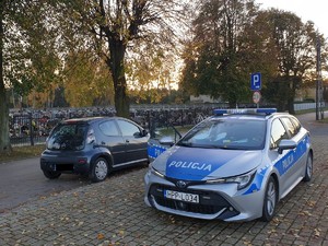 Na zdjęciu radiowóz zaparkowany obok cmentarza- policjanci kontrolują pojazd i jego kierowcę.