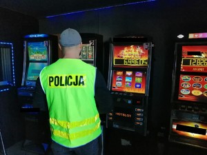 Na zdjęciu policjant w kamizelce odblaskowej z napisem Policja stojący na tle zabezpieczonych automatów do gier hazardowych.