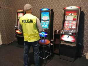Na zdjęciu widoczny policjant stojący przy zabezpieczonym automacie do gier hazardowych.