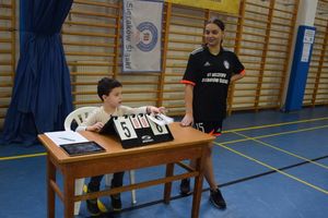 Na zdjęciu widoczny chłopiec siedzący przy stole, stojąca kobieta na hali sportowej.