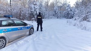 Na zdjęciu policjant stojący obok radiowozu, w tle ośnieżony las.