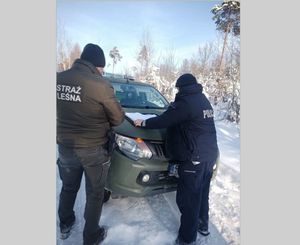 Na zdjęciu widoczny policjant oraz strażnik leśny, w tle samochód oraz ośnieżony krajobraz, z tyłu widoczne drzewa pokryte śniegiem.