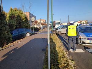 Na zdjęciu policjant ruchu drogowego pracujący przy wypadku, widoczny rozbity samochód osobowy oraz radiowóz policyjny.