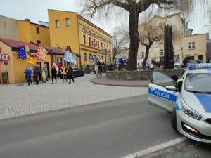 Na zdjęciu widoczny policyjny radiowóz blokujący drogę, wysiadający z niego policjant, orasz przemarsz Orszaku Trzech Króli, mężczyźni przebrani za monarchów jadący na koniach oraz zebrani na uroczystości wierni.