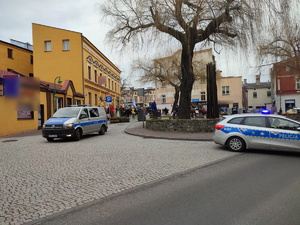 Na zdjęciu widoczne dwa radiowozy, jeden z nich blokuje drogę, w tle schodzący z lublinieckiego rynku wierni biorący udział we procesji.