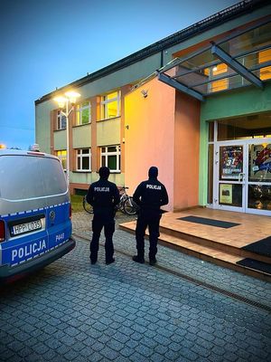 Na zdjęciu widoczni policjanci stojący przy radiowozie i obserwujący Miejski Dom Kultury.