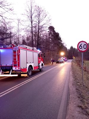 Na zdjęciu widoczny radiowóz z włączonymi sygnałami uprzywilejowania, za nim ustawiony jest samochód straży pożarnej, za nim strażacy  oraz rozbity samochód osobowy.