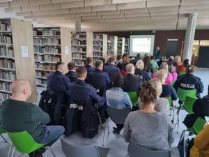 Na zdjęciu widoczni policjanci wraz z słuchaczami siedzący w sali lublinieckiej biblioteki. Z przodu sali prelegentka oraz widoczna prezentacja multimedialna.