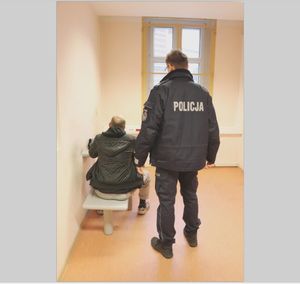 Na zdjęciu umundurowany policjant stojący przy siedzącym zatrzymanym mężczyźnie w pomieszczeniu dla osób zatrzymanych.