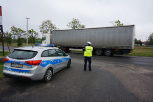 na zdjęciu widoczny jest oznakowany radiowóz , umundurowany policjant w kamizelce odblaskowej z napisem Policja. Policjant stoi przy jezdni , którą przejeżdża samochód ciężarowy