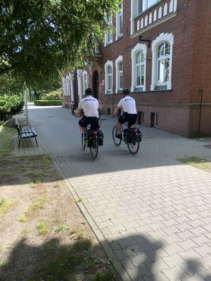 na zdjęciu widoczny jest patrol rowerowy składający się z dwóch policjantów w umundurowaniu , biała koszulka polo z napisem policja , oraz krótkie spodenki w czarnym kolorze