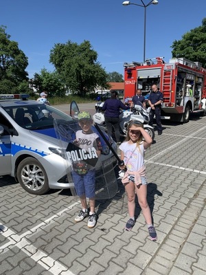 na zdjęciu widoczny jest chłopiec i dziewczynka za ich plecami widoczny jest oznakowany radiowóz