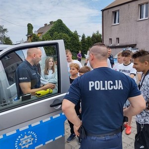 na zdjęciu widoczny jest tyłem policjant w koszulce z napisem policja oraz widoczny jest radiowóz oraz dzieci