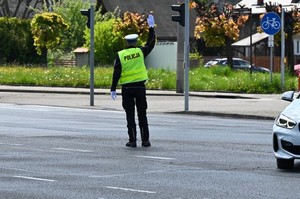 na zdjęciu widoczny jest policjant ruchu drogowego stojący na skrzyżowaniu z podniesiona dłonią,  kierujący ruchem