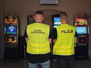 na zdjęciu widoczni są funkcjonariusze Krajowej Administracji Skarbowej oraz Policji w kamizelkach stojący tyłem przed nimi widoczne są automaty do gier losowych