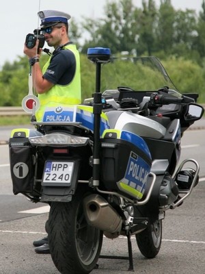 na zdjęciu widoczny jest umundurowany policjant stojący przy motocyklu i trzymający w ręku urządzanie do pomiaru prędkości