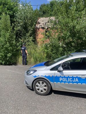 na zdjęciu widoczny jest policjant  sprawdzający pustostan obok stoi oznakowany radiowóz