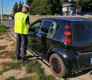 na zdjęciu widoczny jest umundurowany policjant w kamizelce odblaskowej kontrolujący samochód osobowy koloru czarnego