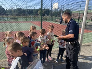 na zdjęciu widoczny jest umundurowany policjant rozdający dzieciom stojącym wokół niego odblaskowe opaski na rękę