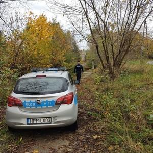 na zdjęciu widoczny jest oznakowany radiowóz oraz umundurowany policjant idący w stronę opuszczonych budynków wśród drzew