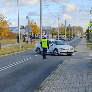 na zdjęciu widoczny jest umundurowany policjant stający na ulicy i kierujący ruchem , widoczne jest również auto osobowe koloru białego