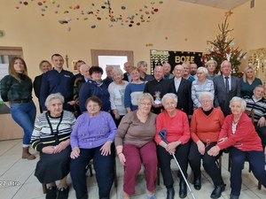 na zdjęciu widoczna jest grupa seniorów siedzących na krzesłach , a część stojąca pozujących do wspólnego zdjęci wśród nich widoczny ksiądz w sutannie