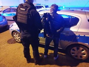 na zdjęciu widoczny jest policjant umundurowany stojący obok oznakowanego radiowozu z osoba zatrzymana